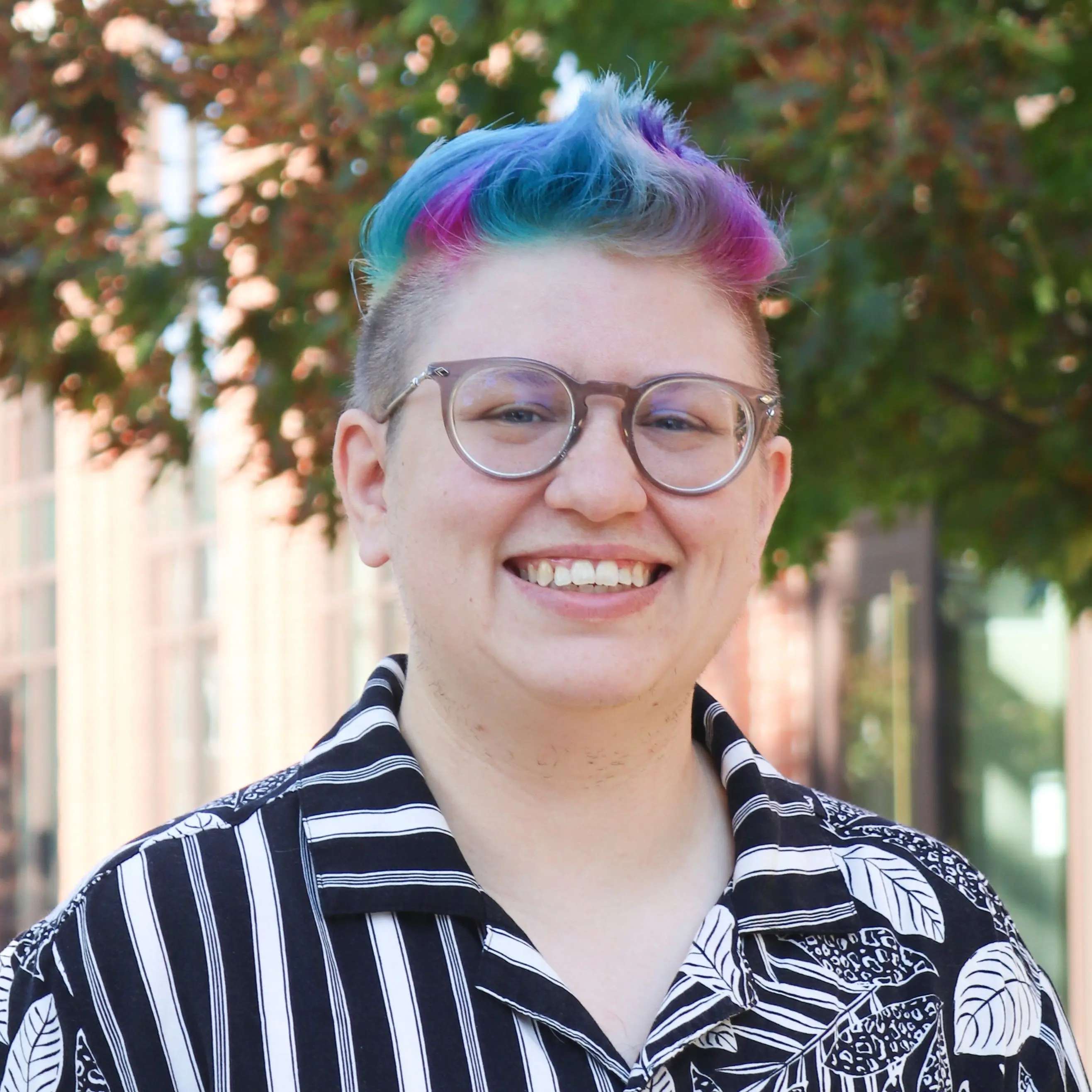 A headshot of Andy. They have short, multicolored hair, large, round glasses, and wear a black-and-white patterned shirt. They are outdoors with trees and a brick building in the background.