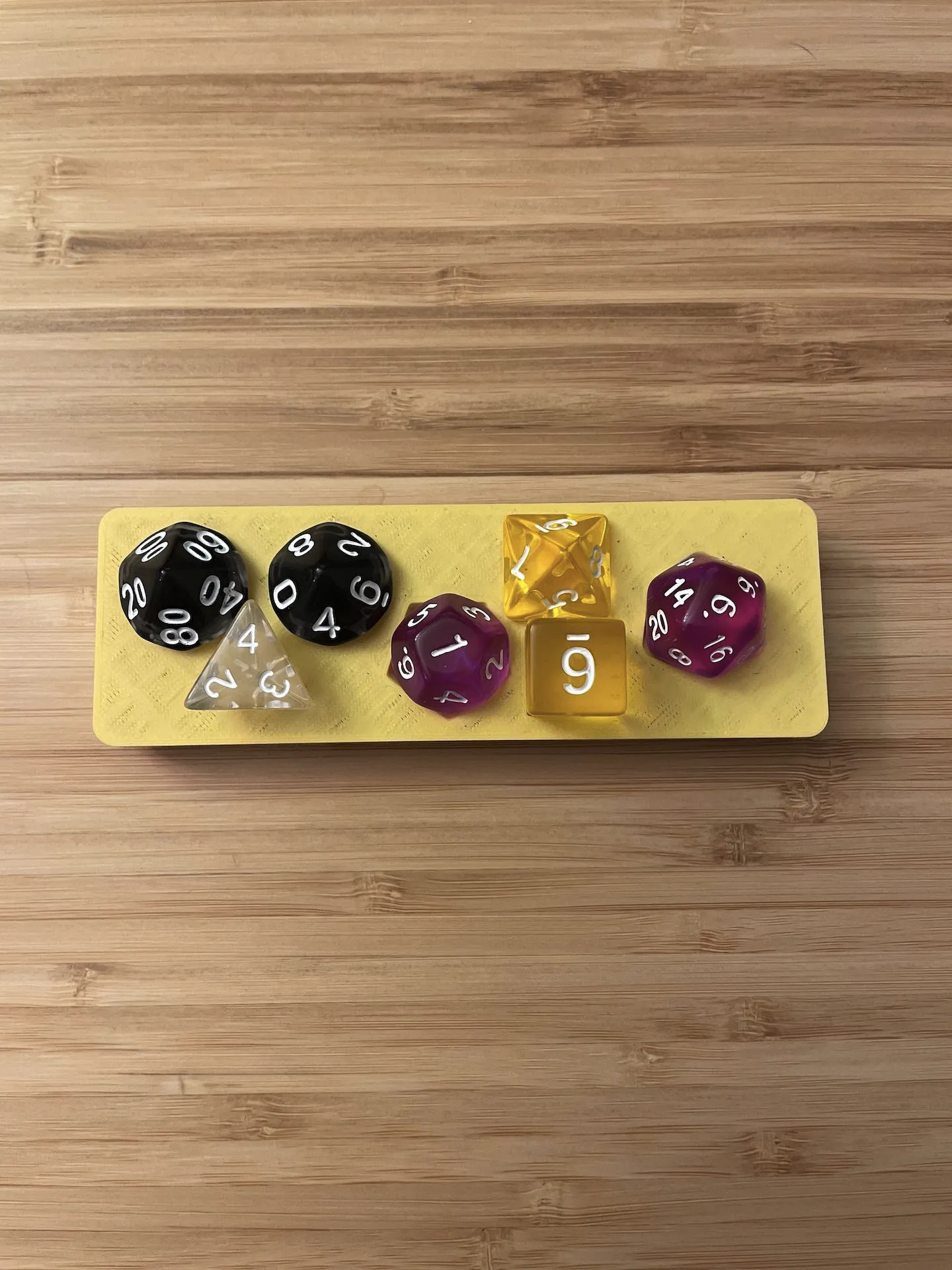 7 dice in assorted sizes, shapes and colors arranged neatly on a matte yellow 3D printed bin. The bin is on a bamboo desk.