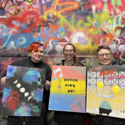 Three people holding their spray-painted canvases in front of a graffiti-covered wall.