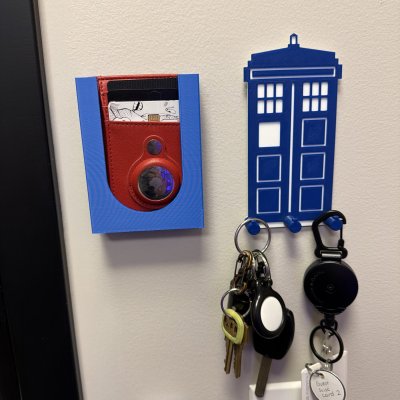 A 3D-printed blue TARDIS key hook holding keys and a badge reel, next to a red and blue printed AirTag holder on the wall.