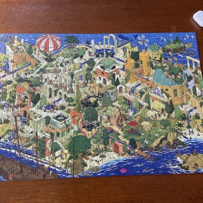 A finished jigsaw puzzle of an illustrated fantasy city by the sea, on a wooden table.