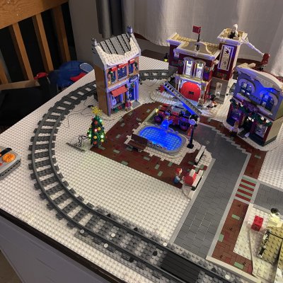 A LEGO winter village on a table: a train track curves around a town square with a lit Christmas tree, a blue ice rink and colorful storefronts.
