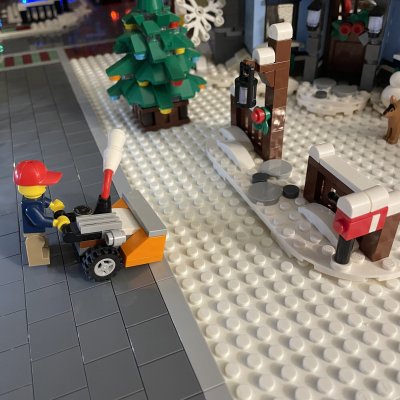 A LEGO minifigure in a red cap clearing snow with a yellow snowblower next to a Christmas tree.