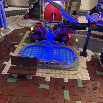 A red LEGO fire truck with its ladder raised outside the fire station, beside a glowing blue skating pond.