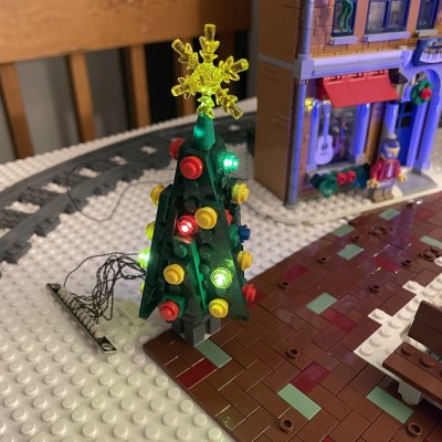 A lit LEGO Christmas tree with colored lights and a gold star on a brick-paved square in front of a music shop.