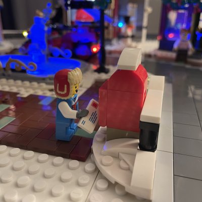 A LEGO mail carrier dropping a letter into a red mailbox, with lit buildings blurred behind.