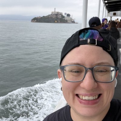 Andy grinning in a backwards cap on the ferry, with the wake behind them and Alcatraz Island in the distance.