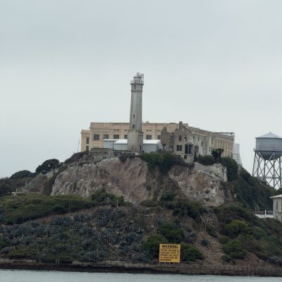 Alcatraz Island from the water: the lighthouse and cellhouse on top of the rocky cliffs under a grey sky.