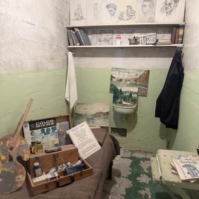 A small cell staged with a paint box, a palette, sketches pinned above a shelf of books, and a coat hanging by the sink.
