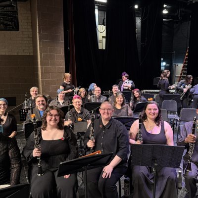 A concert band dressed in black sitting on stage with their instruments, smiling at the camera.