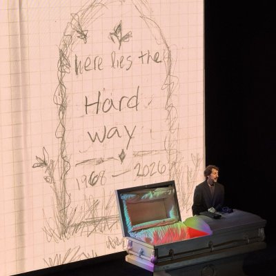 Dave Kiss on a dark stage beside an open coffin, in front of a giant sketched tombstone that reads “Here lies the Hard way, 1968–2026.”