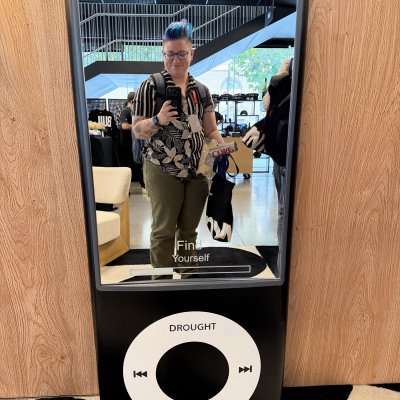 Andy's reflection in the mirrored screen of a giant iPod nano prop that reads “Now Living.”
