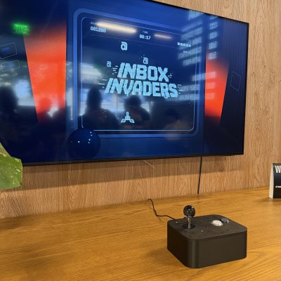 An arcade-style Inbox Invaders game on a wall-mounted TV, with a joystick on the wooden counter below.