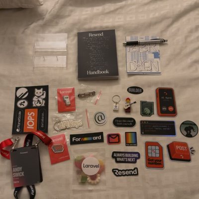 Conference swag spread out on a hotel bed: stickers, pins, a notebook, a lanyard, and Resend and Laravel goodies.
